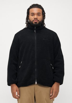 Man with dreadlocks wearing black zip-up fleece jacket and beige pants, standing against plain white background.