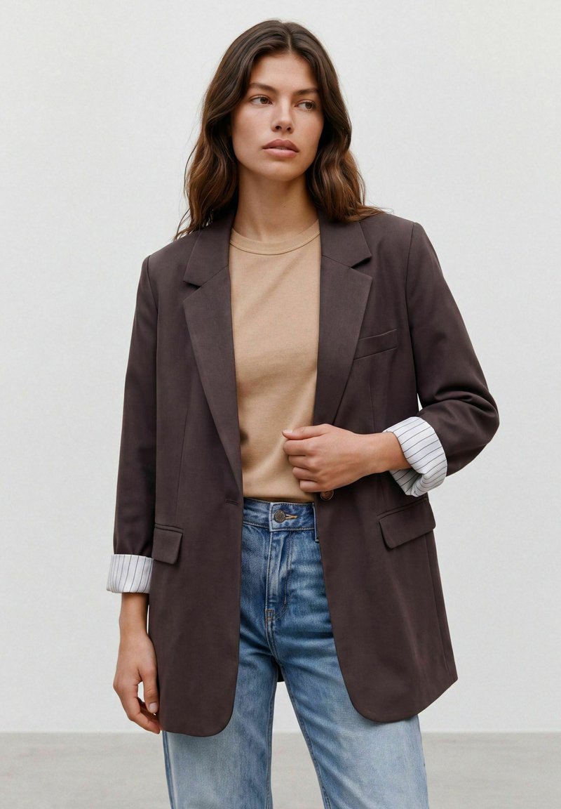 Femme aux cheveux bruns ondulés portant un blazer marron foncé sur une chemise beige et un jean bleu clair, debout devant un fond clair uni.