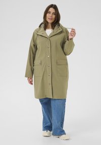 Femme portant un imperméable vert olive à boutons pression, jean large bleu, baskets blanches, tenant le cordon de la capuche sur un fond uni.