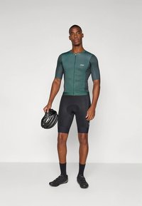Ciclista masculino de pie sosteniendo un casco negro, vistiendo una camiseta de ciclismo verde, pantalones cortos acolchados negros, calcetines negros y zapatos de ciclismo negros, fondo simple.