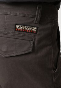 Mörkgrå byxor med en texturerad tygkonstruktion, som har en bakficka och en framträdande svart och röd broderad etikett.