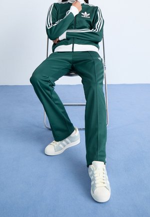 Personne assise sur une chaise en métal portant un survêtement vert Adidas et des baskets blanches Adidas sur un sol recouvert de moquette bleue.