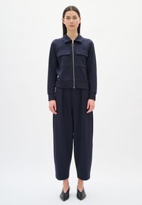 Giacca blu navy con zip e due tasche sul petto, abbinata a pantaloni blu navy a vita alta e tacchi neri a punta, contro uno sfondo bianco.