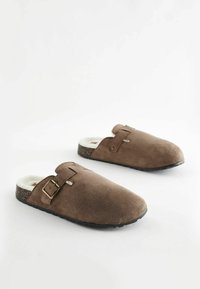 Hnědé semišové pantofle s huňatým vnitřkem, s přezkou a texturovanou tmavou podrážkou. Zaoblený design se otevřenou patou.