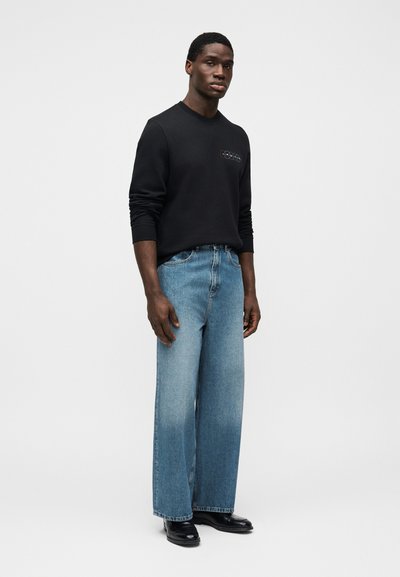 Μαύρο φούτερ με στρογγυλή λαιμόκοψη, μαλακή υφή, συνδυασμένο με ανοιχτό μπλε τζιν παντελόνι φαρδιάς γραμμής. Το μοντέλο στέκεται μπροστά σε ουδέτερο φόντο.