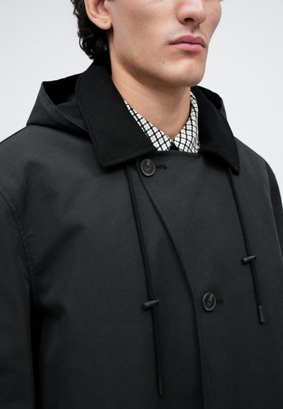 Man wearing a black hooded coat with button details over a patterned black and white collared shirt.