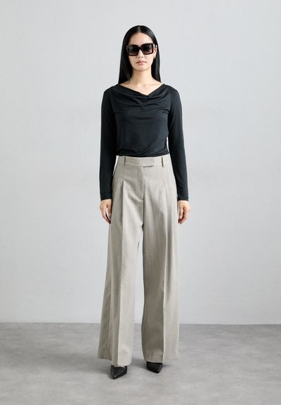 Black long-sleeve top with a draped neckline, paired with high-waisted, wide-leg gray trousers and black pointed-toe heels. Large black sunglasses.