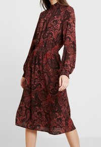 Vestido de estampado paisley rojo con mangas largas, cuello alto y cintura fruncida. Hecho de una tela ligera, cae por debajo de la rodilla.