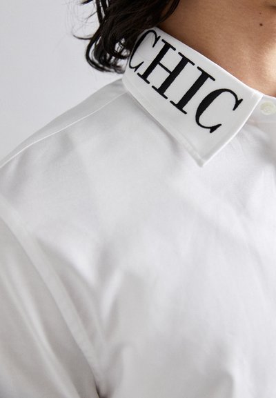 White collared shirt featuring a prominent black embroidered "CHIC" on the collar. Smooth texture with subtle sheen. Classic button-up design.