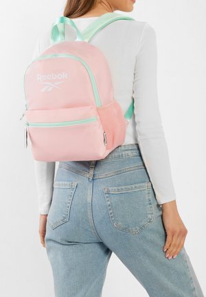 Kobieta ubrana w jasnoróżowy plecak Reebok z miętowozielonymi paskami, białą bluzkę z długim rękawem i jasnoniebieskie dżinsy, pozująca tyłem do kamery.