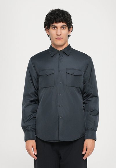 Chemise noire à manches longues en matériau lisse et léger. Dotée d'une fermeture à boutons sur le devant, de deux poches poitrine et d'un col classique.