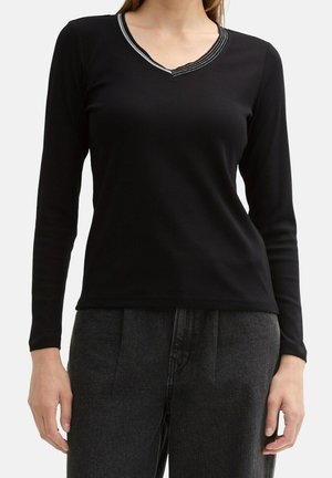 Kobieta w czarnej bluzce z długim rękawem i dekoltem w serek z białym przeszyciem, w połączeniu z ciemnoszarymi spodniami z wysokim stanem.