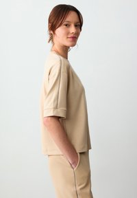 Beżowa bluzka z krótkim rękawem, wykonana z miękkiego materiału, o luźnym kroju, z okrągłym dekoltem i widocznymi szarymi szwami na ramionach.