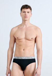 Μυώδης νεαρός άνδρας που φοράει μαύρα εσώρουχα με γκρι μέση, στέκεται μπροστά σε απλό φωτεινό φόντο, κοιτάζοντας προς τα εμπρός.