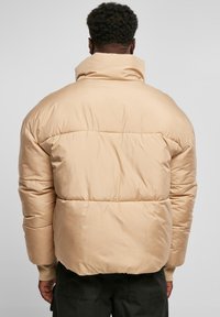 Beige dunjakke med høy krage, kviltet design og myk, glatt tekstur. Har elastiske mansjetter og en lett cropped passform.