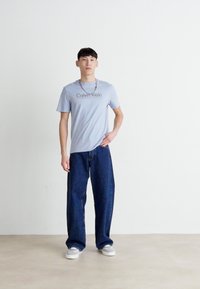 Šviesiai mėlynas medvilninis marškinėliai su "Calvin Klein" logotipu, derinami su tamsiai mėlynomis džinsinio audinio plačiomis kelnėmis ir pilkais šlepetėmis, stovintis ant neutralios fono.
