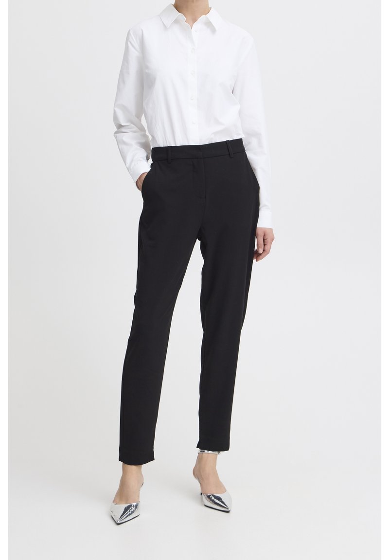 Personne portant une chemise blanche boutonnée, un pantalon noir sur mesure et des talons pointus argentés sur un fond clair uni.