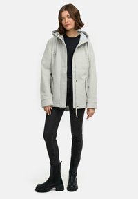 Une femme portant une veste à capuche gris clair par-dessus un t-shirt noir, un jean skinny noir et des bottes chunky noires, se tenant dans une posture détendue.