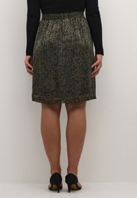 Jupe plissée noire avec un motif floral dans des tons de vert et d'or. Taille élastique, longueur genou, associée à des chaussures à talons noires.