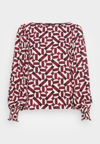 Blouse à manches longues avec un motif géométrique rouge, noir et blanc. Le tissu a une texture lisse avec des poignets élastiqués et un large décolleté.
