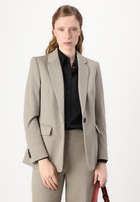 Bež jakna s krojem i reverima, teksturirana tkanina, dva prednja džepa i zatvaranje na jedan gumb. Nosi se preko crne košulje na dugmad.