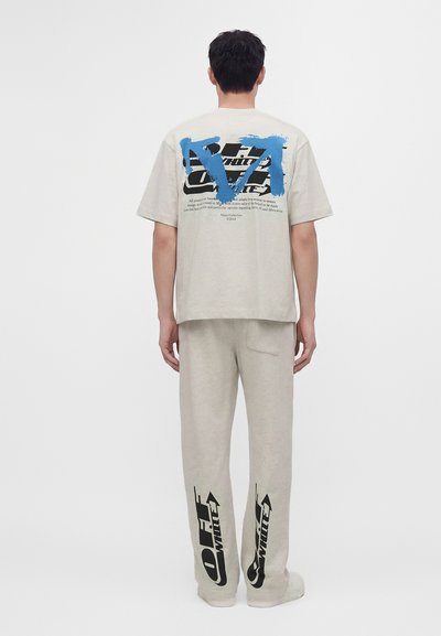 Άνδρας που φοράει μπεζ παντελόνια Off-White με μαύρα λογότυπα στα πόδια και μπεζ μπλουζάκι με μαύρο και μπλε γραφικό κείμενο στην πλάτη.