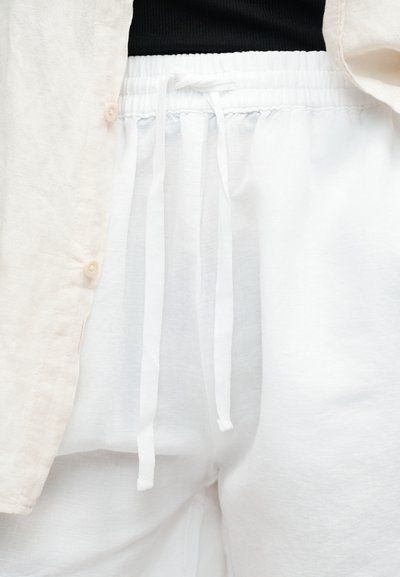 Κοντινό πλάνο ατόμου που φοράει λευκό παντελόνι με κορδόνι, μπεζ πουκάμισο με κουμπιά και μαύρο τοπ μέσα στο παντελόνι.