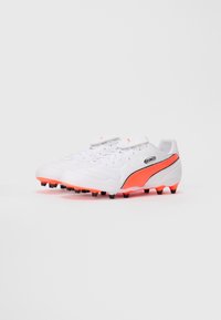 Pari valkoisia Puma King -jalkapallokenkiä, joissa on oranssi raita ja pohjan nastat, vinosti vasemmalle, yksinkertaisella valkoisella taustalla.