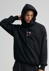 Sudadera de algodón negra con una capucha amplia, bolsillo tipo canguro y texto impreso en rosa. Presenta puños acanalados y un parche de logo en la manga.