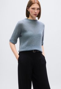 Ανοιχτό μπλε πουλόβερ με κοντά μανίκια σε απαλό υλικό, στρογγυλή λαιμόκοψη, φορεμένο με μαύρο ψηλόμεσο παντελόνι και λεπτή ζώνη, χέρια στις τσέπες.
