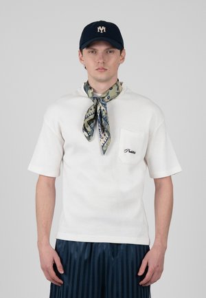 Homme portant une chemise blanche à manches courtes avec une poche devant, un foulard à motifs autour du cou, un pantalon rayé bleu foncé et une casquette bleu marine avec logo.