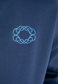 Tela azul con diseño simétrico de bucles bordados en azul claro, centrado en la superficie.