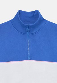 Blå kvart-zip genser med en ribbestrikket krage. Har en rosa stripe over brystet over et hvitt panel. Myk, teksturert bomullsstoff.