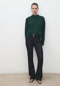 Maglione a maglia verde scuro con colletto alto, abbinato a jeans neri a zampa d'elefante. Accessoriato con una cintura nera e sandali con cinturini.