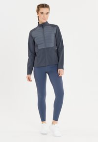 Kvinna som står i en blå vadderad jacka, marinblå leggings och vita sneakers mot en enkel vit bakgrund.