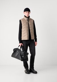Beige quiltet vest, svart turtleneck, mørke jeans, svart skinn duffelbag og svarte sko. Har en glidelås foran og runde håndtak.