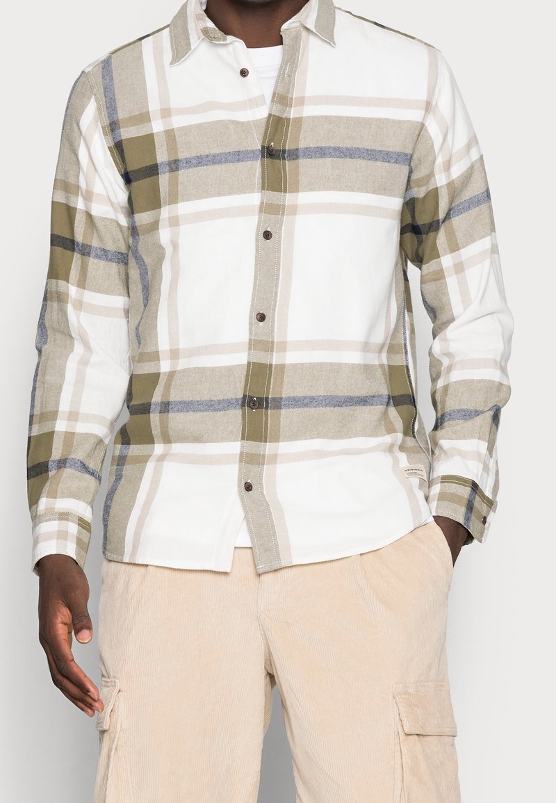 Man i en beige och vit rutig skjorta med knäppning över en vit topp, ihop med ljusbeige kordflorsbyxor med fickor, med händerna i fickorna.