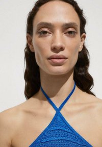 Blue halter top made of a textured, shimmering material. Features a triangular shape and crisscross straps around the neck. Smooth skin tones.