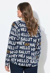 Námořnická mikina s dlouhým rukávem, zdobená bílým a oranžovým textem s pozdravy v různých jazycích, texturovaný tisk s opotřebovaným efektem.