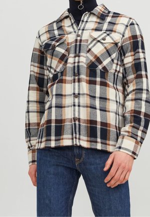 Chemise à carreaux boutonnée en crème, marine et marron, avec deux poches poitrine, un col évasé et une texture à motifs. Portée avec un jean foncé.