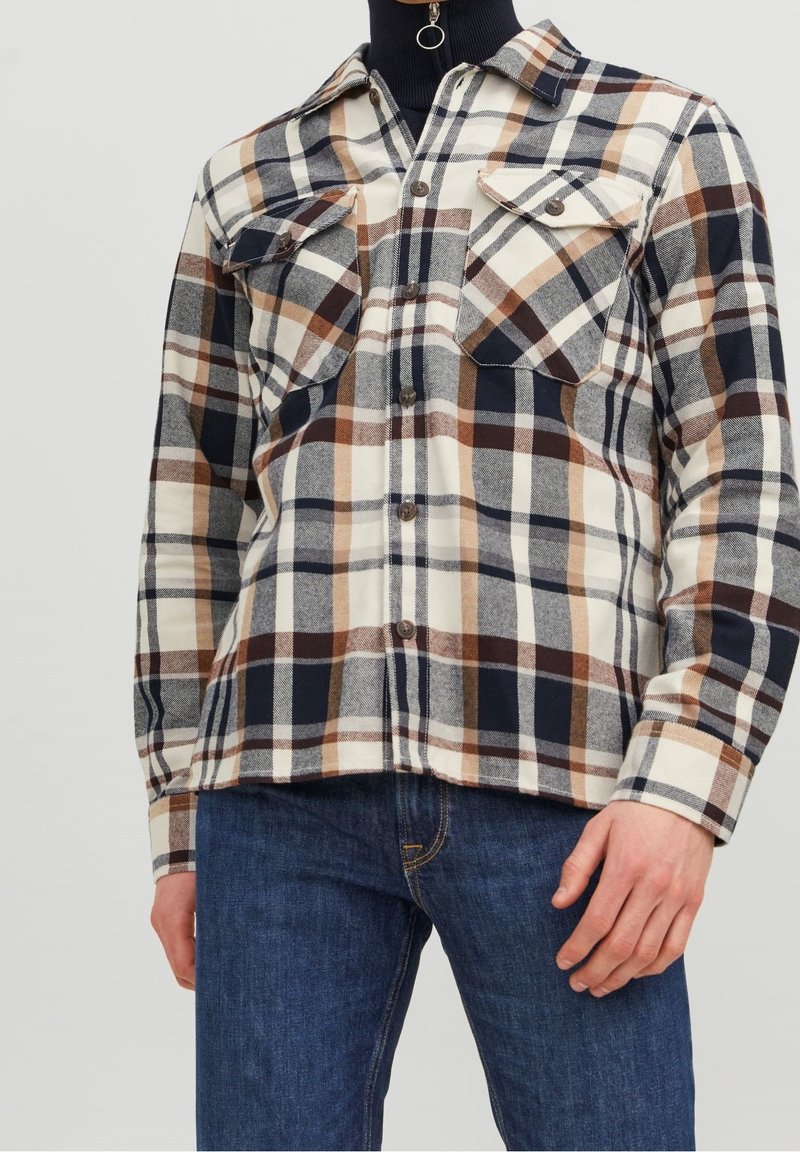 Camicia a scacchi in crema, blu navy e marrone, con due tasche sul petto, colletto strutturato e texture a motivi. Indossata con jeans scuri.