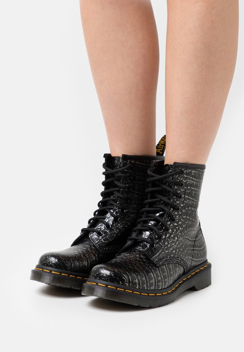 Bottes noires en cuir avec une texture crocodile, bout rond, lacets sur le devant, surpiqûres jaunes en accent, et semelle épaisse.