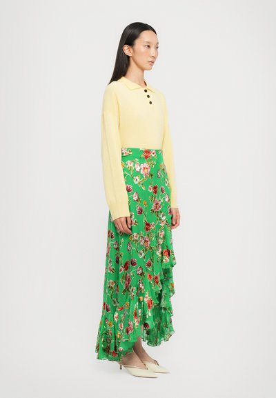 Polo giallo a maniche lunghe con dettagli a bottoni abbinato a una gonna maxi verde floreale con orlo arricciato e vivaci motivi multicolore.