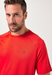 Rød atletisk t-shirt lavet af glat, let materiale. Har en rund halsudskæring, korte raglanærmer og et lille blåt logo på brystet.