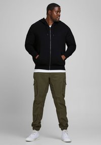 Black zip-up hoodie with front pockets, olive green cargo pants, and white sneakers. The outfit features a casual, relaxed fit.