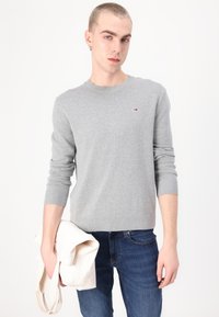 Grijze gebreide trui met een ronde hals en geribde manchetten, voorzien van een klein logo op de borst, gecombineerd met blauwe jeans en een lichte totebag.