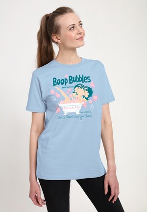 Lyseblåt t-shirt med et grafisk design af et boblebad med teksten "Boop Bubbles Mild formula." Model i sorte bukser.