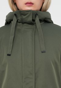 Veste vert olive avec un col montant et des cordons de serrage. Présente une texture lisse et des accessoires en métal près des extrémités des cordons.