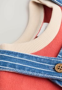 Koraliowy sweter z prążkowanym kremowym kołnierzem, dżinsowy ogrodnik z białymi paskami i dekoracyjnym guzikiem. Teksturowany materiał i warstwowy design.