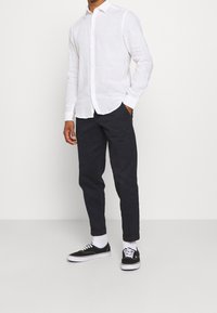 Muž v bílém košili s dlouhým rukávem a knoflíky, černých nohavicích s manžetami, bílých ponožkách a černých šněrovacích teniskách stojí na jednobarevném pozadí.